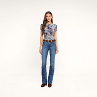 Une femme se tient debout devant un fond beige. Elle porte le t-shirt ajusté Ressi à motif paisley bleu nuit, associé à un jean bleu, une ceinture marron et des chaussures marron. Elle a de longs cheveux bruns ondulés et regarde droit devant elle avec une expression neutre.