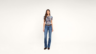 A woman stands against a plain white background, wearing a blue patterned top, blue flared jeans, a brown belt, and brown shoes. She faces forward with a relaxed expression.