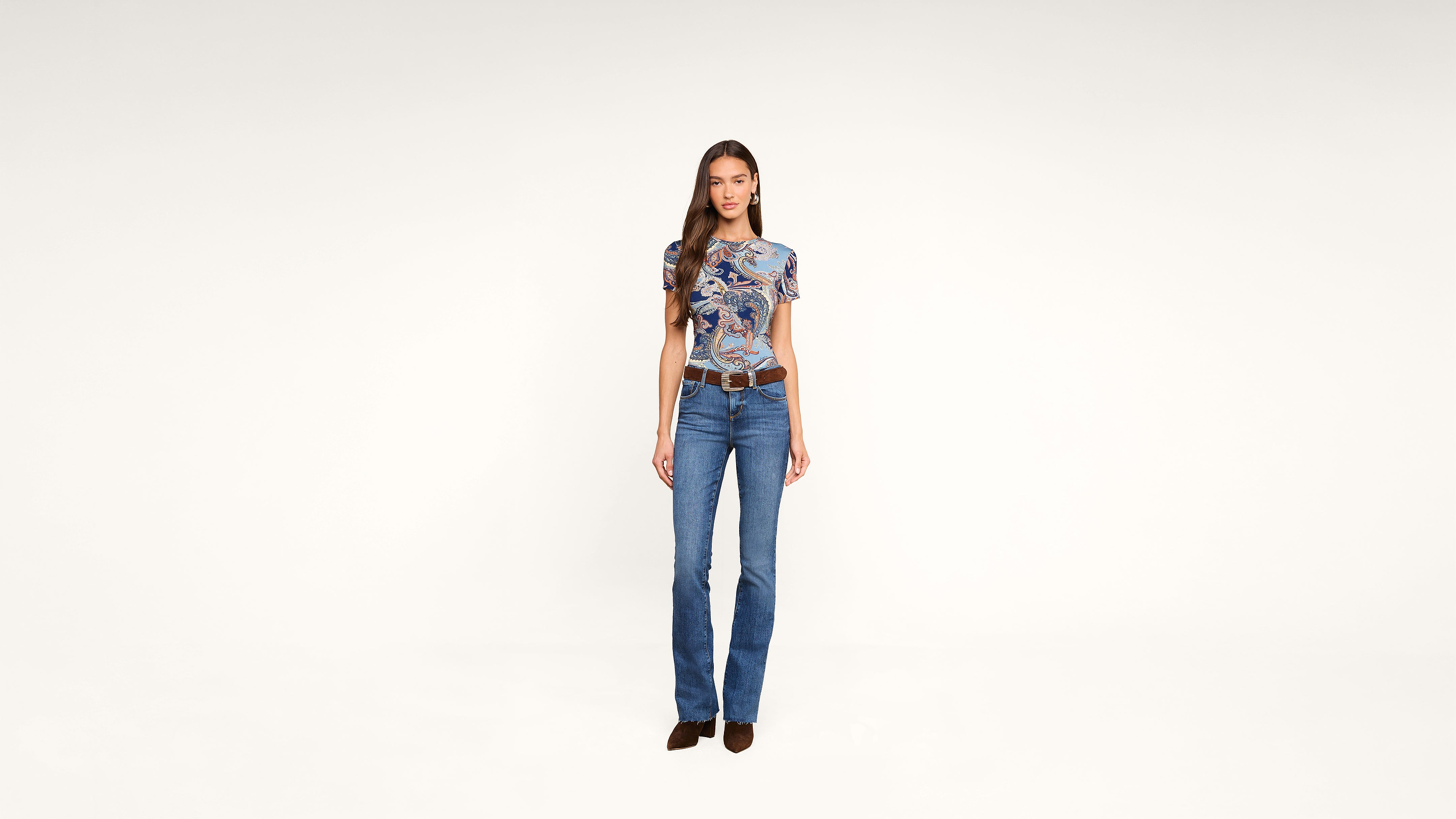 A woman stands against a plain white background, wearing a blue patterned top, blue flared jeans, a brown belt, and brown shoes. She faces forward with a relaxed expression.