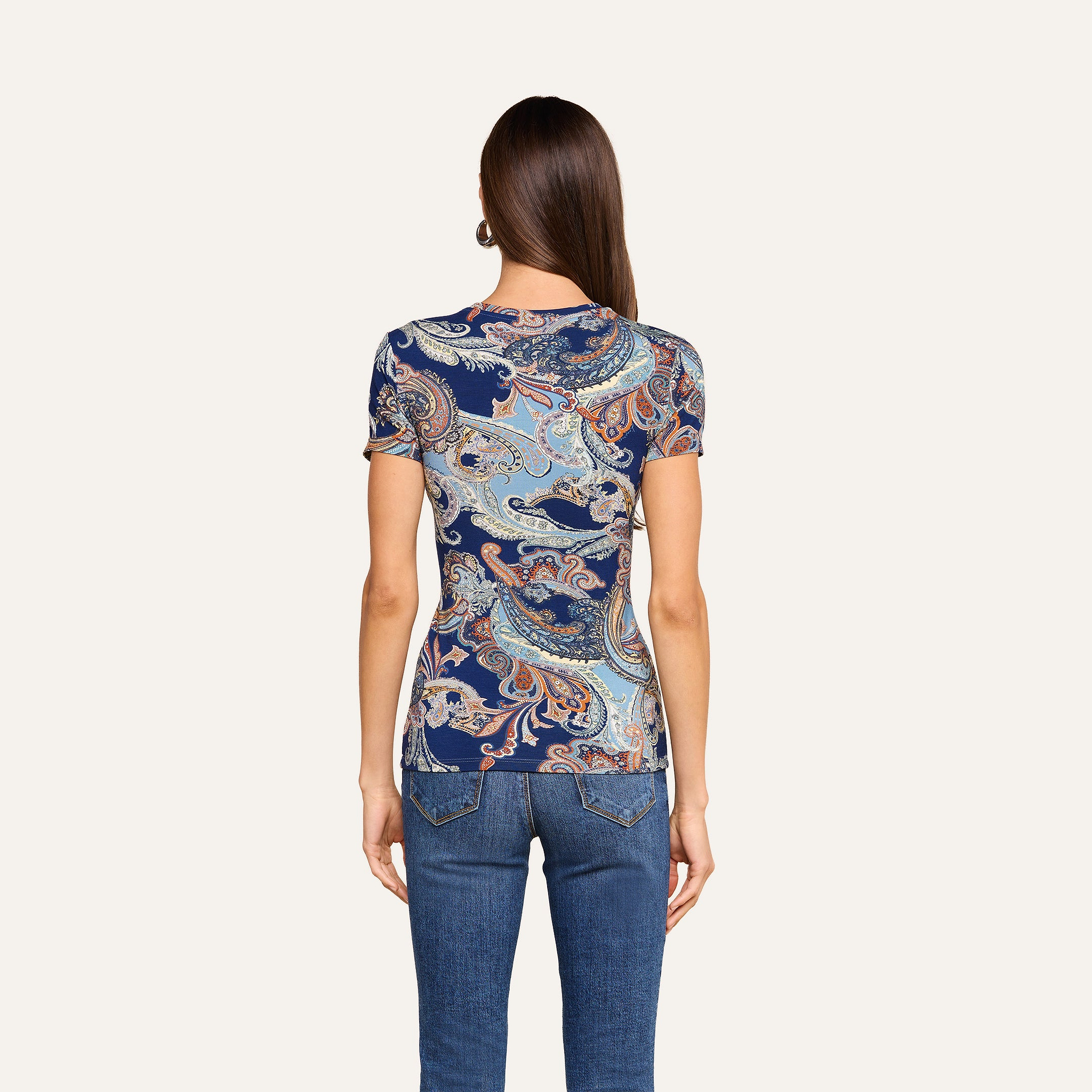 On voit de dos une femme aux longs cheveux bruns et raides, vêtue dun t-shirt ajusté Ressi à imprimé paisley bleu nuit et dun jean, se tenant debout devant un fond beige uni.