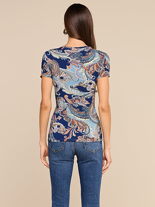 A woman with long, straight brown hair is seen from behind wearing the Ressi Fitted Tee in a midnight blue paisley print and a pair of jeans, standing against a plain beige background.
