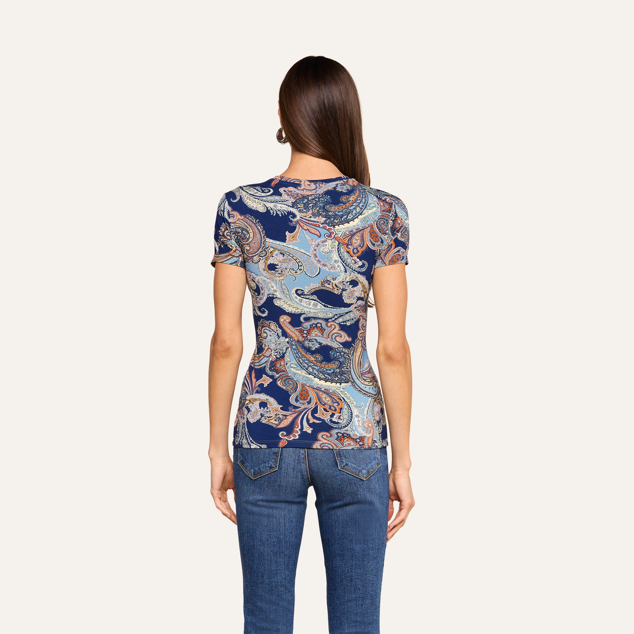 On voit de dos une femme aux longs cheveux bruns et raides, vêtue dun t-shirt ajusté Ressi à imprimé paisley bleu nuit et dun jean, se tenant debout devant un fond beige uni.