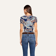 On voit une femme de dos, vêtue du t-shirt ajusté Ressi au motif paisley bleu nuit, quelle porte avec un jean bleu et une ceinture marron, sur un fond beige uni.