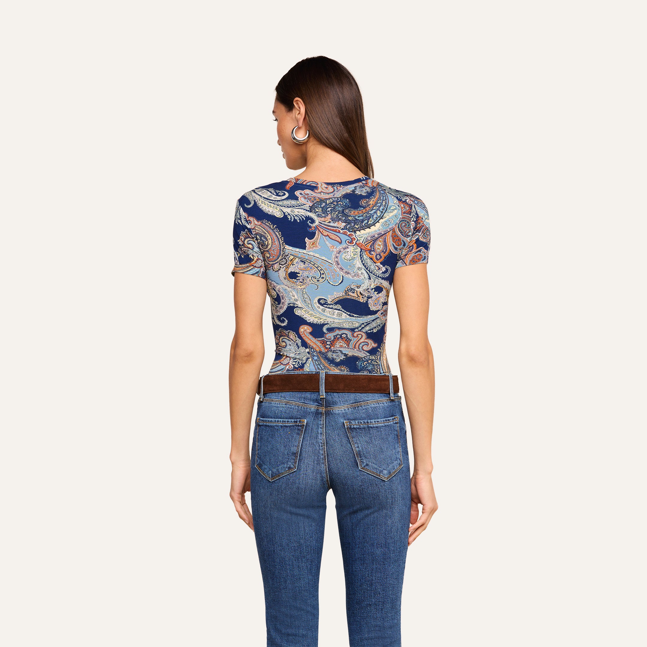On voit une femme de dos, vêtue du t-shirt ajusté Ressi au motif paisley bleu nuit, quelle porte avec un jean bleu et une ceinture marron, sur un fond beige uni.