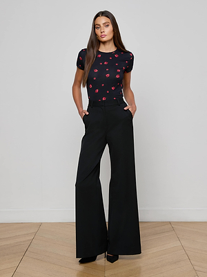 A woman with long brown hair stands against a light-gray wall, wearing the black Ressi Fitted Tee with a small red rose print and high-waisted, wide-leg black trousers. Her hands are in her pockets.