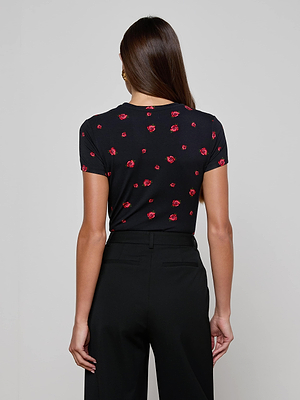 A woman faces away from the camera, wearing a Ressi Fitted Tee in stretch jersey with a bold red rose print and black trousers, set against a plain light gray background.