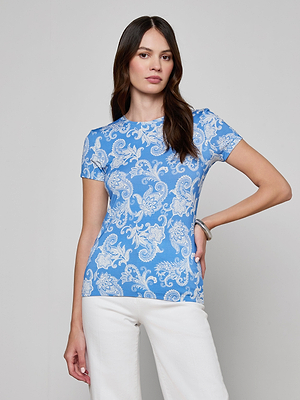 A woman with long brown hair poses confidently against a light background, wearing the bestselling Ressi Fitted Tee in light blue with a white floral design and matching white pants. The slim stretch jersey flatters her figure.