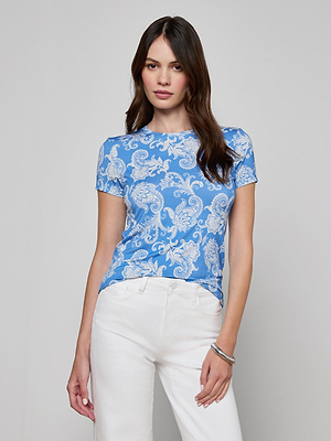 A woman with long brown hair wears the Ressi Fitted Tee, a slim-cut blue and white paisley stretch jersey top, paired with white pants against a plain light gray background.