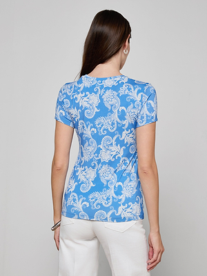 A woman with long brown hair, seen from behind, wears the Ressi Fitted Tee in light blue stretch jersey knit with a white paisley pattern and white pants, standing against a plain light gray background.