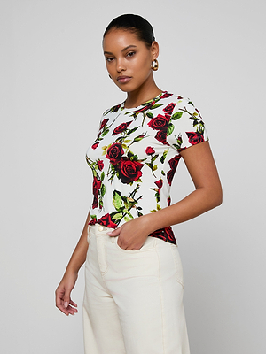 A woman stands against a plain background wearing the bestselling Ressi Fitted Tee in white stretch jersey with a red rose print, cream slim-cut pants, gold hoop earrings, and her hair tied back. One hand is in her pocket and she has a neutral expression.