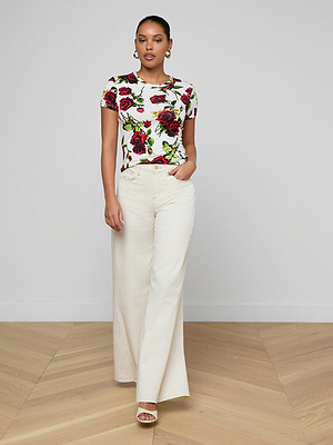 A woman stands indoors in the bestselling Ressi Fitted Tee—a white, short-sleeve stretch jersey top with a red rose print—paired with cream wide-leg pants and open-toed heels against a light wall and wooden floor.