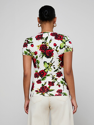 A person with dark hair in a bun and gold hoop earrings models the bestselling Ressi Fitted Tee in white with a red rose pattern and off-white pants, shown from the back against a plain light gray background.