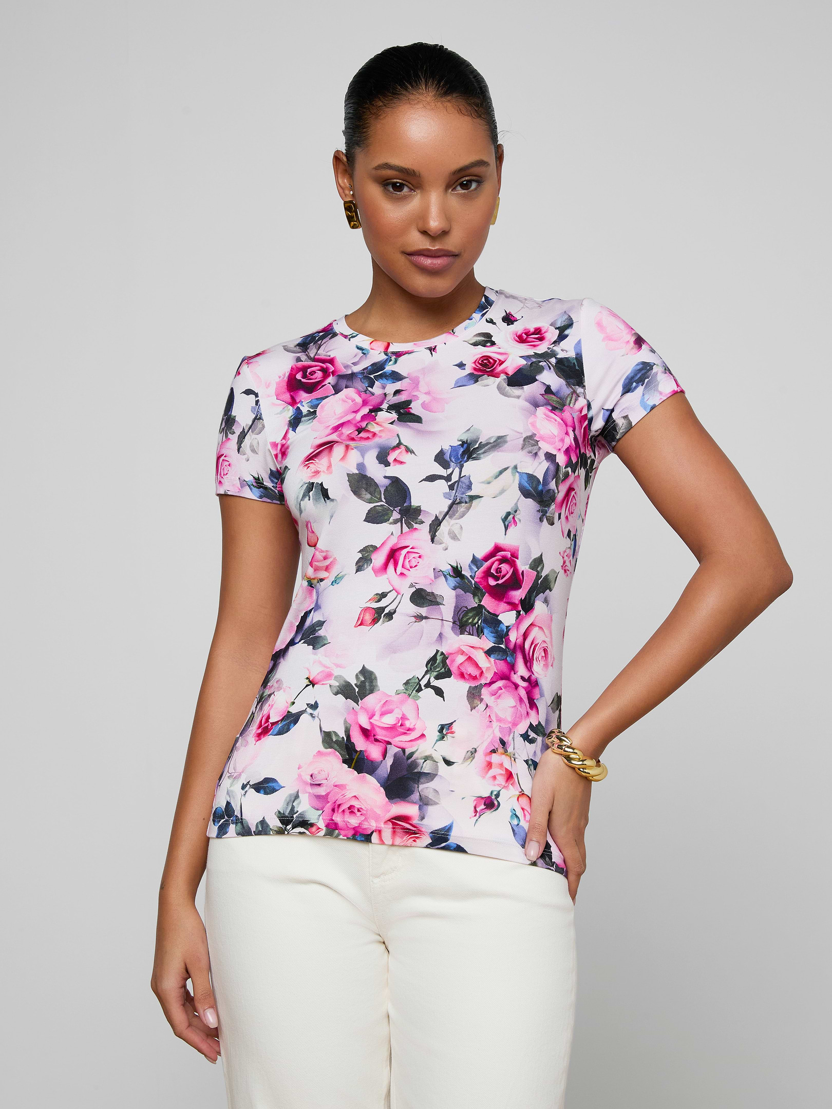A woman poses with one hand on her hip against a plain light background, wearing the Ressi Fitted Tee—a bestselling white short-sleeved top featuring a pink rose and purple floral print—paired with white pants.