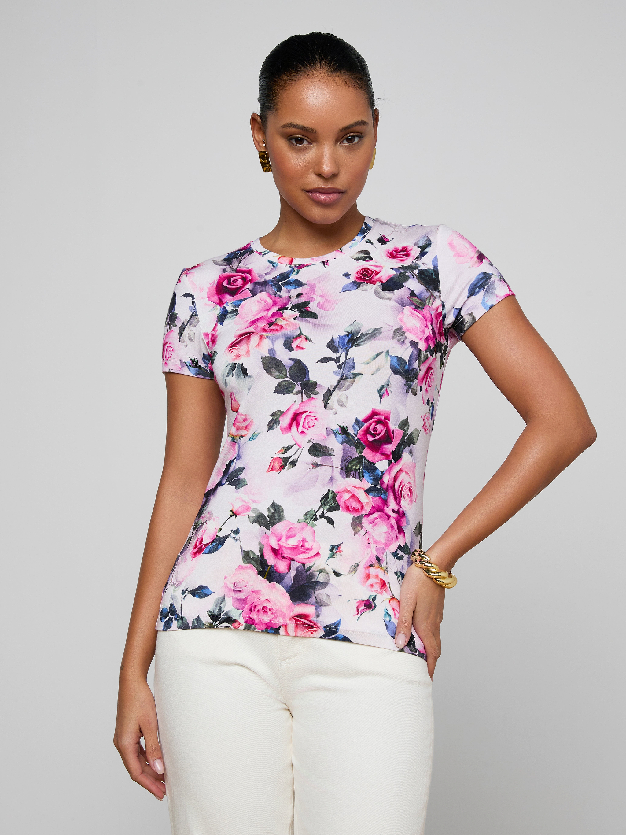 A woman poses confidently in the bestselling Ressi Fitted Tee, a stretch jersey knit with a vibrant pink rose print, paired with white pants against a plain light background.