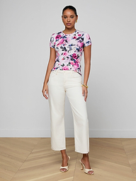 A woman stands indoors wearing the Ressi Fitted Tee in pink rose print stretch jersey, paired with white high-waisted straight-leg pants and beige heeled sandals. She has her hair tied back, accessorizing with gold bracelets against a minimal background.