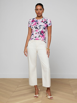 A woman stands indoors wearing the Ressi Fitted Tee in pink rose print stretch jersey, paired with white high-waisted straight-leg pants and beige heeled sandals. She has her hair tied back, accessorizing with gold bracelets against a minimal background.