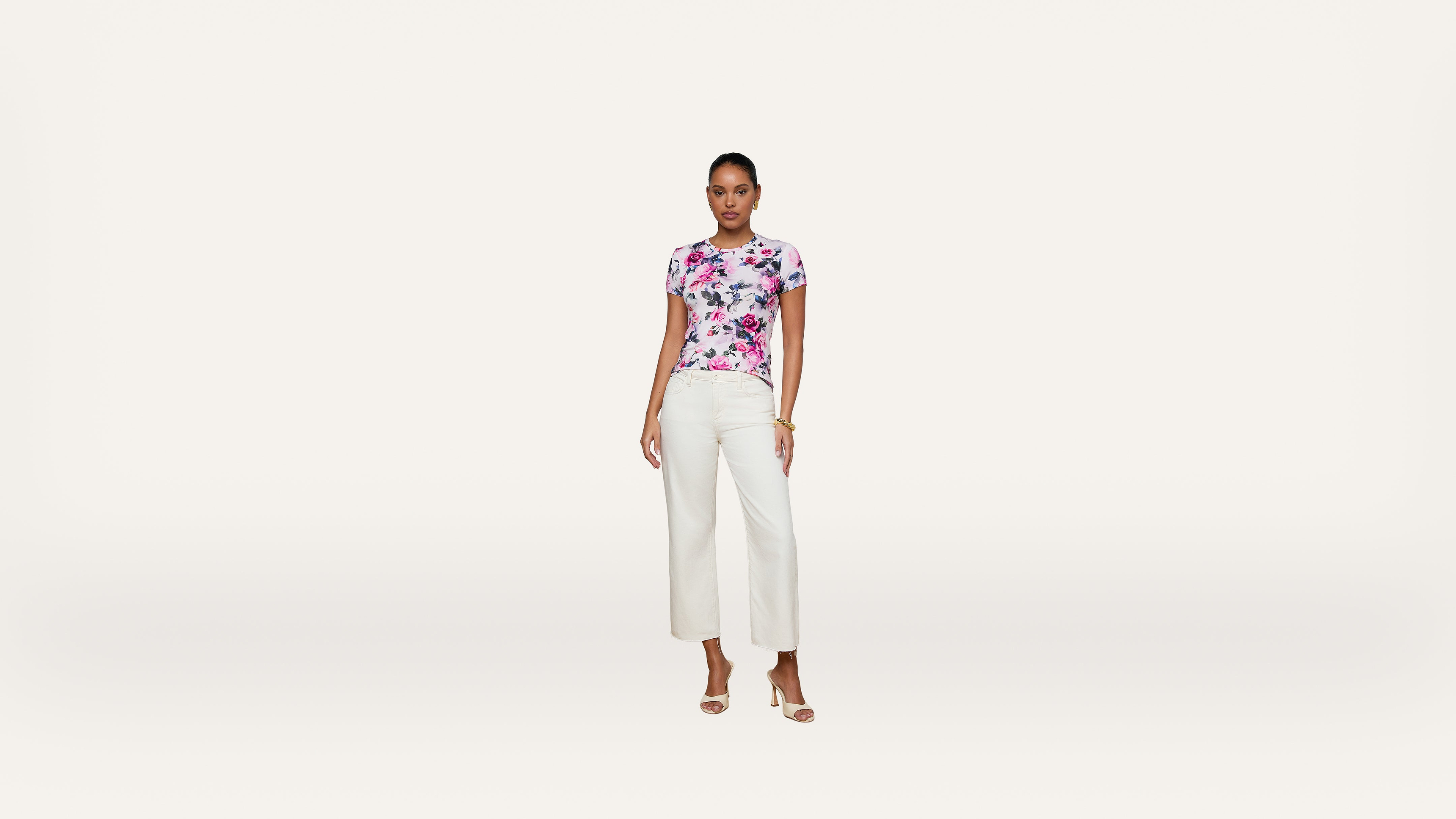 A woman stands against a white background, wearing a floral short-sleeve top, white wide-leg pants, and beige heeled sandals.