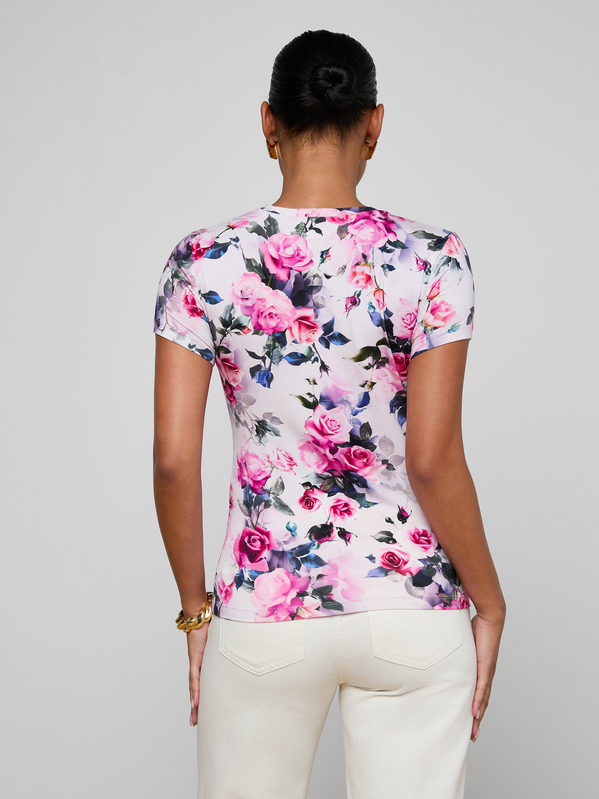 Someone stands facing away, modeling the bestselling Ressi Fitted Tee in stretch jersey knit, featuring a pink rose print, paired with cream-colored pants. Their hair is styled in a bun, set against a plain light gray background.