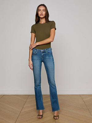 A woman stands indoors against a plain wall, arms crossed and relaxed, wearing the L'AGENCE Ressi Micro-Rib Tee in olive green, paired with blue jeans and brown open-toe heels.