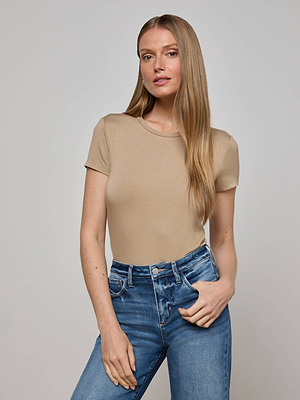 A woman with long, straight blonde hair wears the L'AGENCE Ressi Fitted Tee and blue jeans. She stands against a plain light gray background with one hand in her pocket, gazing confidently at the camera.