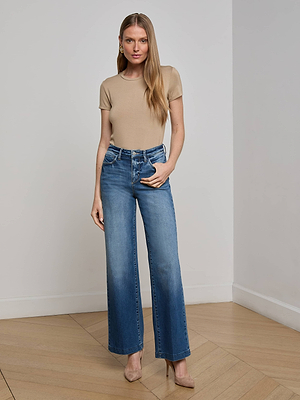 A woman with long blonde hair wears the L'AGENCE Ressi Fitted Tee, high-waisted wide-leg blue jeans, and beige heels as she stands indoors on a wooden floor against a plain white wall, posing with one hand in her pocket.
