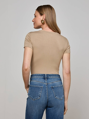 A woman with long, light brown hair wears gold earrings, blue high-waisted jeans, and the L'AGENCE Ressi Fitted Tee as she stands facing away from the camera against a plain light grey background.