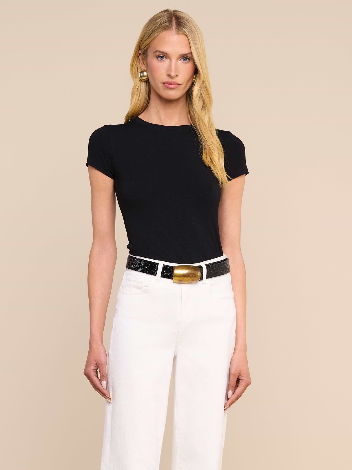 A woman with long blonde hair wears the Ressi Fitted Tee in black, paired with white pants, a black and gold belt, and gold earrings, standing against a plain light background.