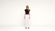 A woman wearing sunglasses, a black t-shirt, white high-waisted pants, and black heels stands against a plain white background, facing forward.