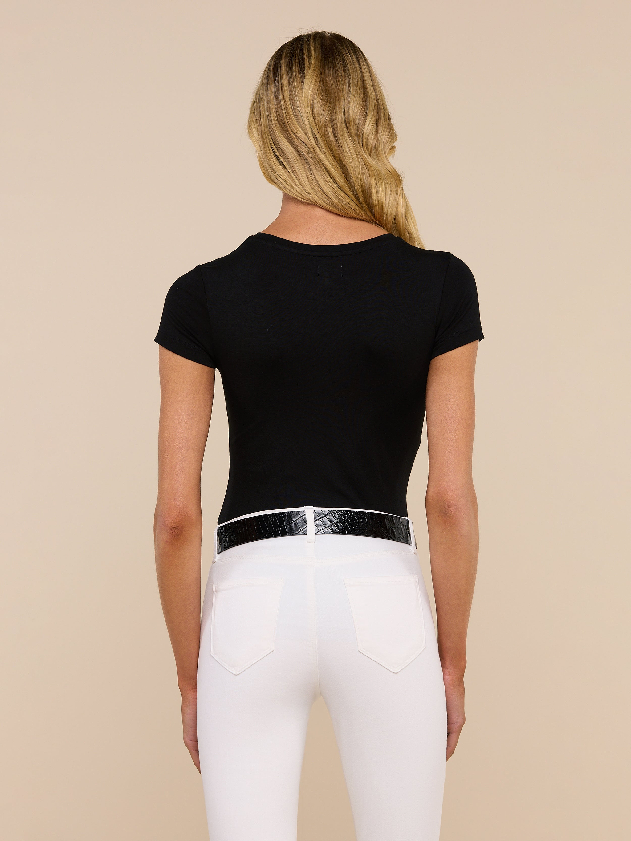 A woman with long blonde hair, wearing the Ressi Fitted Tee in black and white pants, stands facing away from the camera against a plain light background.