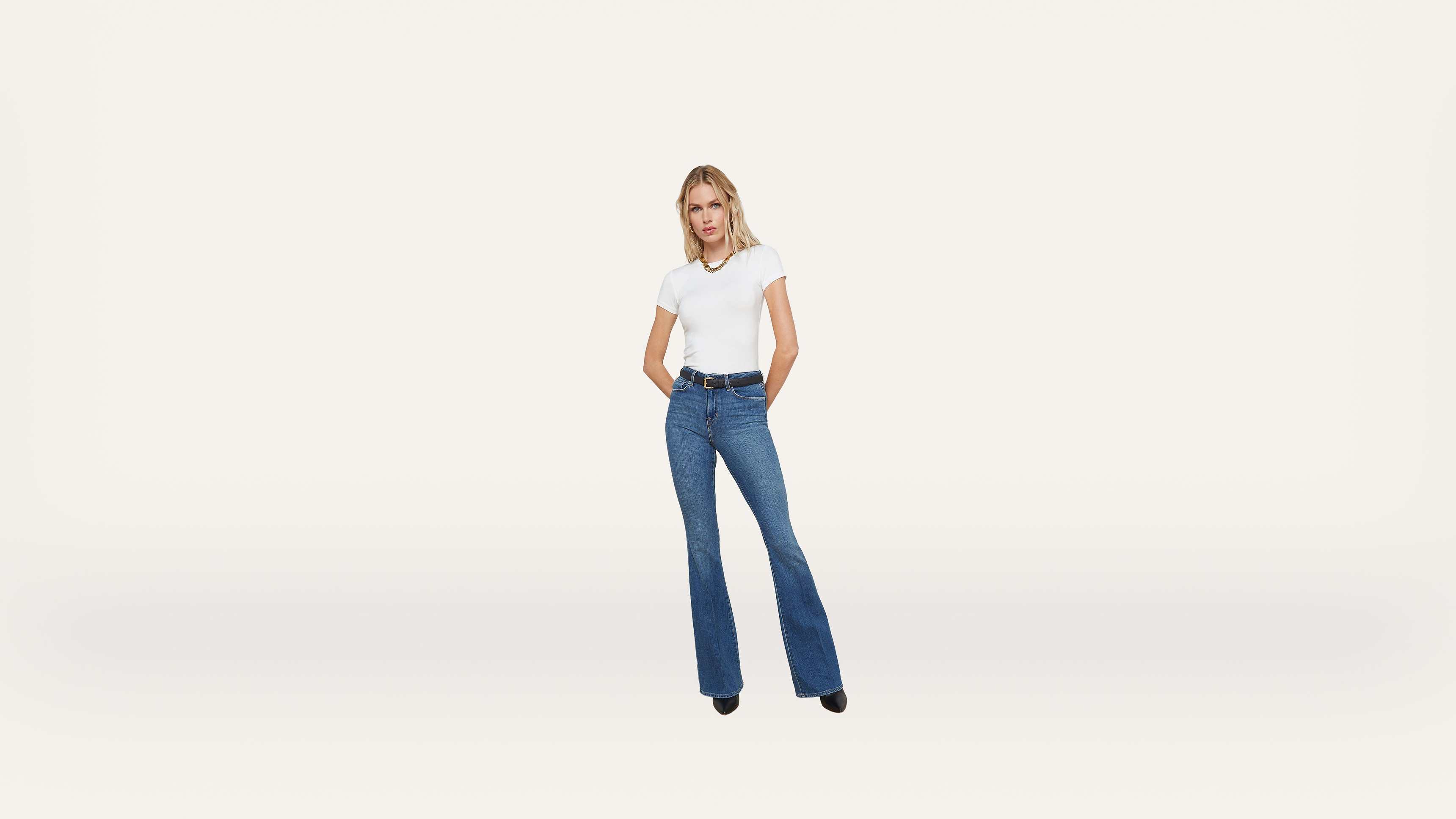 A woman stands against a plain white background, wearing a white t-shirt, blue high-waisted flare jeans, and black shoes. She has her hands in her pockets and is looking straight ahead.