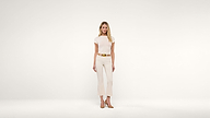 A woman stands against a plain white background, wearing a short-sleeved cream top, cream pants, a tan belt, and tan pointed shoes. She has long blonde hair and a relaxed, neutral expression.