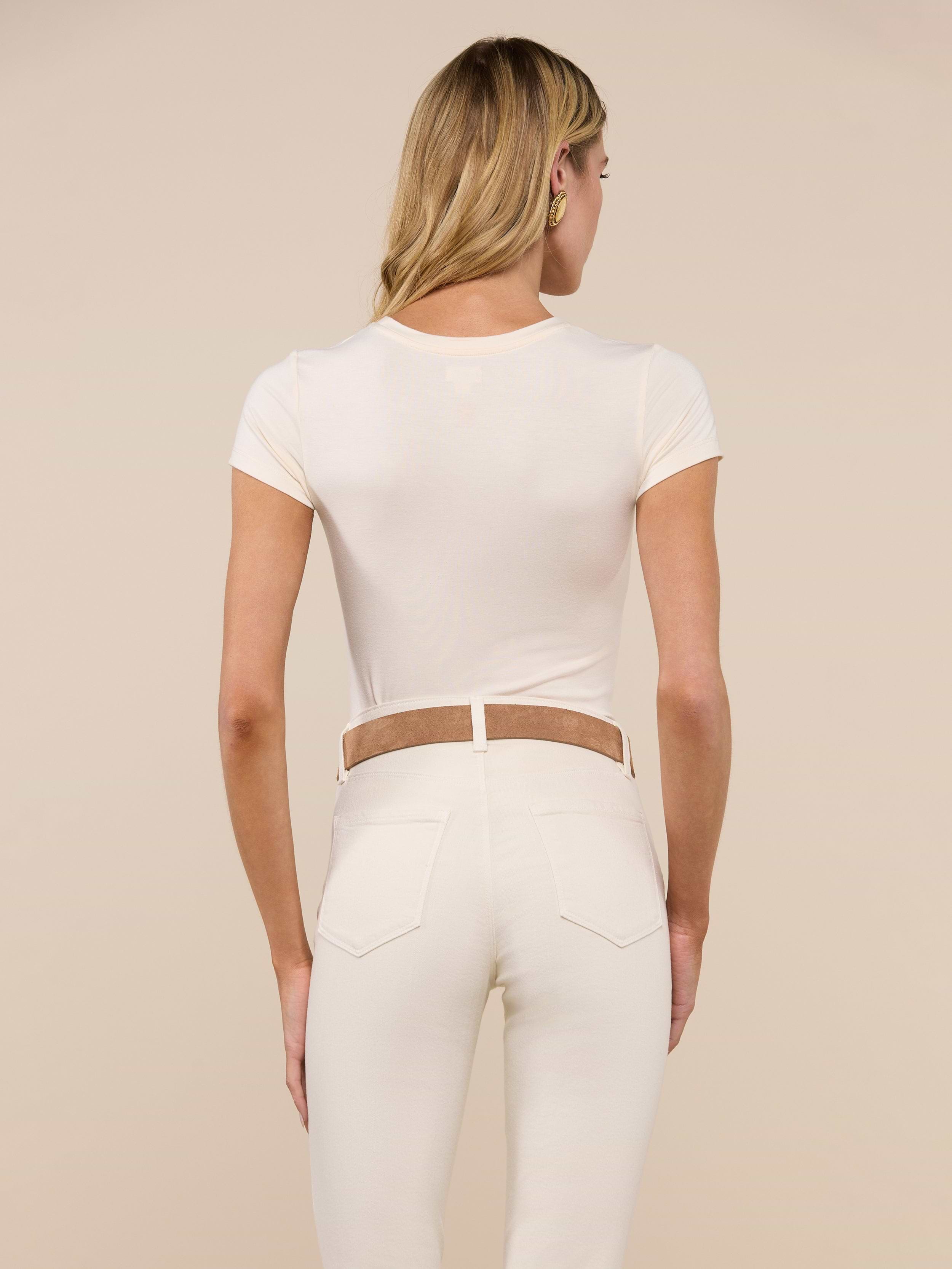 A woman with long blonde hair, wearing the Ressi Fitted Tee in vintage white and high-waisted cream pants with a tan belt, is standing with her back to the camera against a plain background.