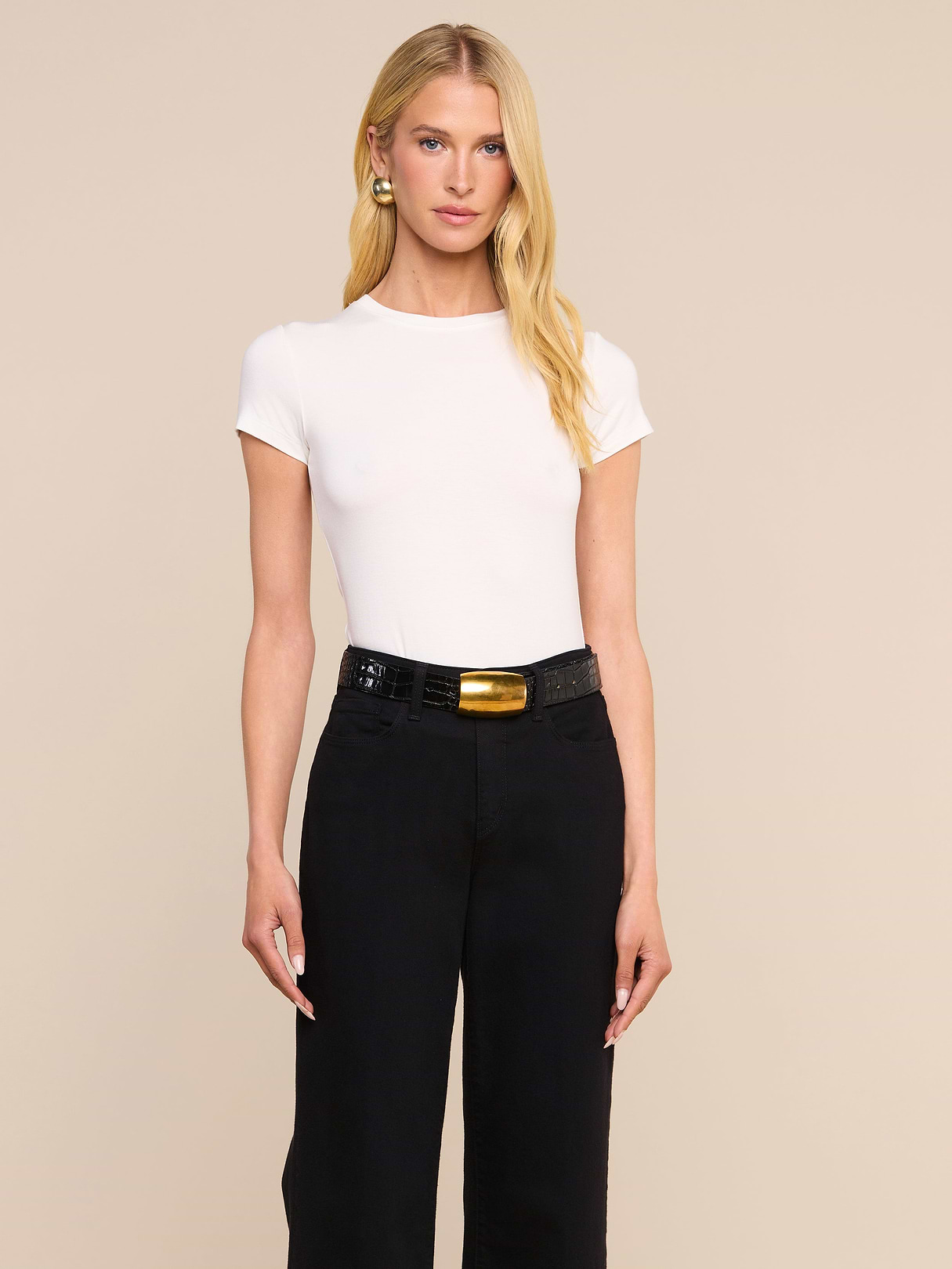 A woman with long blonde hair wears the form-fitting Ressi Fitted Tee in white, tucked into high-waisted black pants with a black belt; she stands against a plain light background.