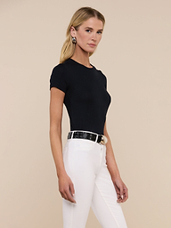 A woman stands against a plain background wearing the black Ressi Micro-Rib Tee styled with white pants, a black belt, and large earrings. She has long blonde hair and a neutral expression.