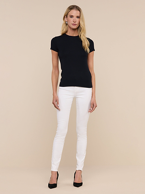 A woman with long blonde hair and a neutral expression stands against a beige background, wearing the Ressi Micro-Rib Tee in black, paired with white skinny jeans and black heels.