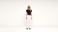 A woman in sunglasses, a black t-shirt, white pants, and black heels stands against a plain white background, facing forward with arms at her sides.