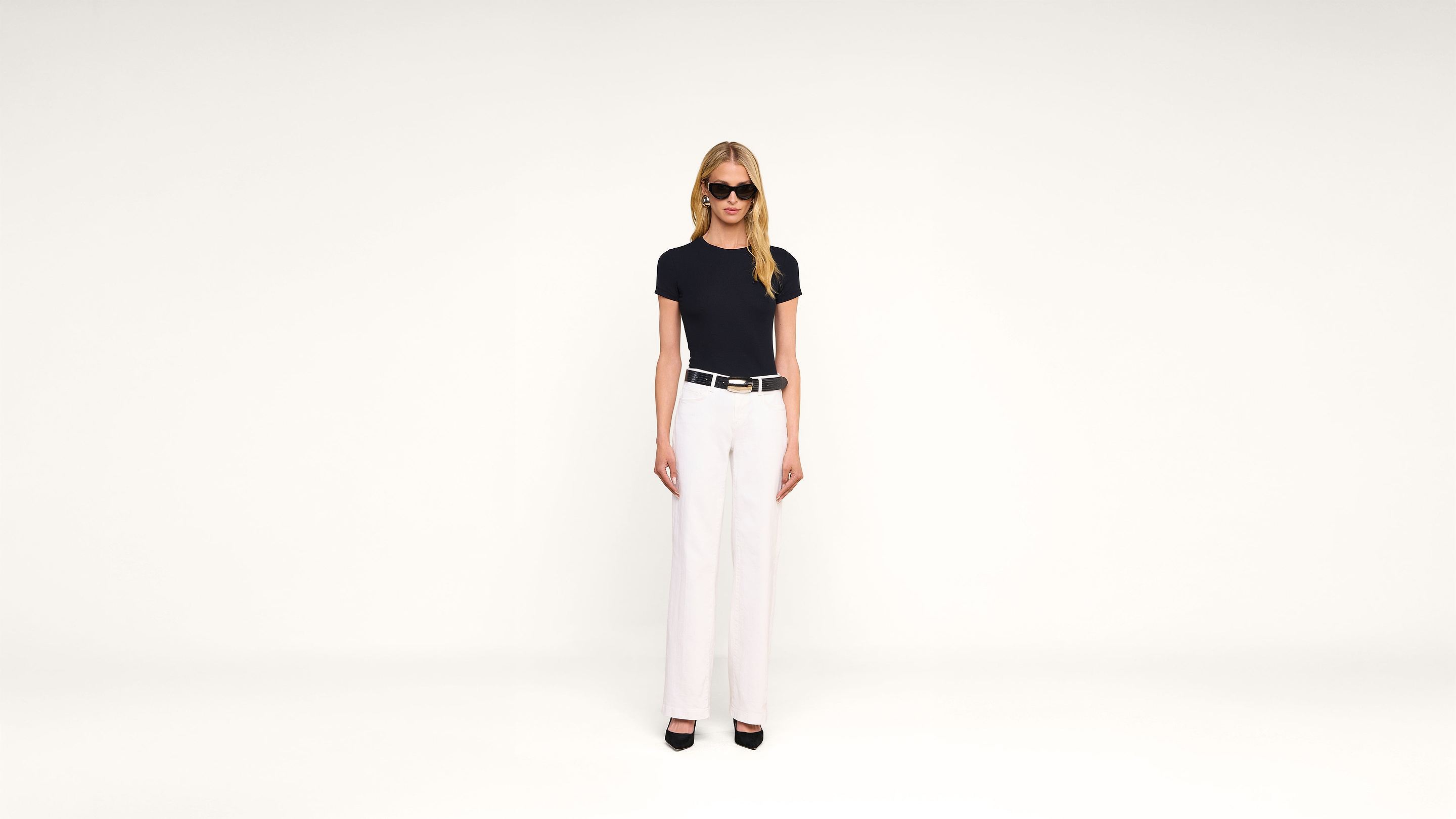 A woman in sunglasses, a black t-shirt, white pants, and black heels stands against a plain white background, facing forward with arms at her sides.