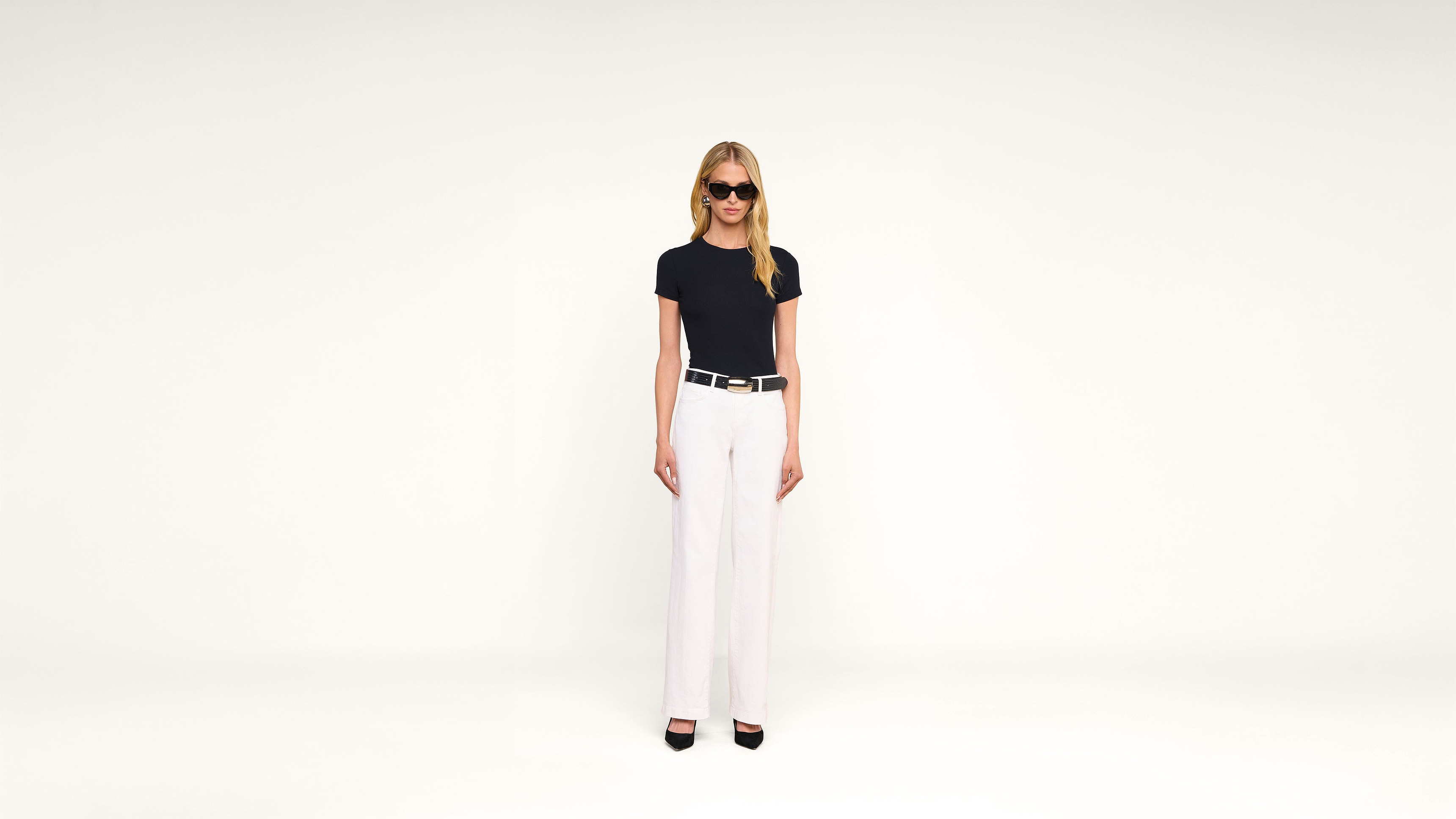 A woman in sunglasses, a black t-shirt, white pants, and black heels stands against a plain white background, facing forward with arms at her sides.