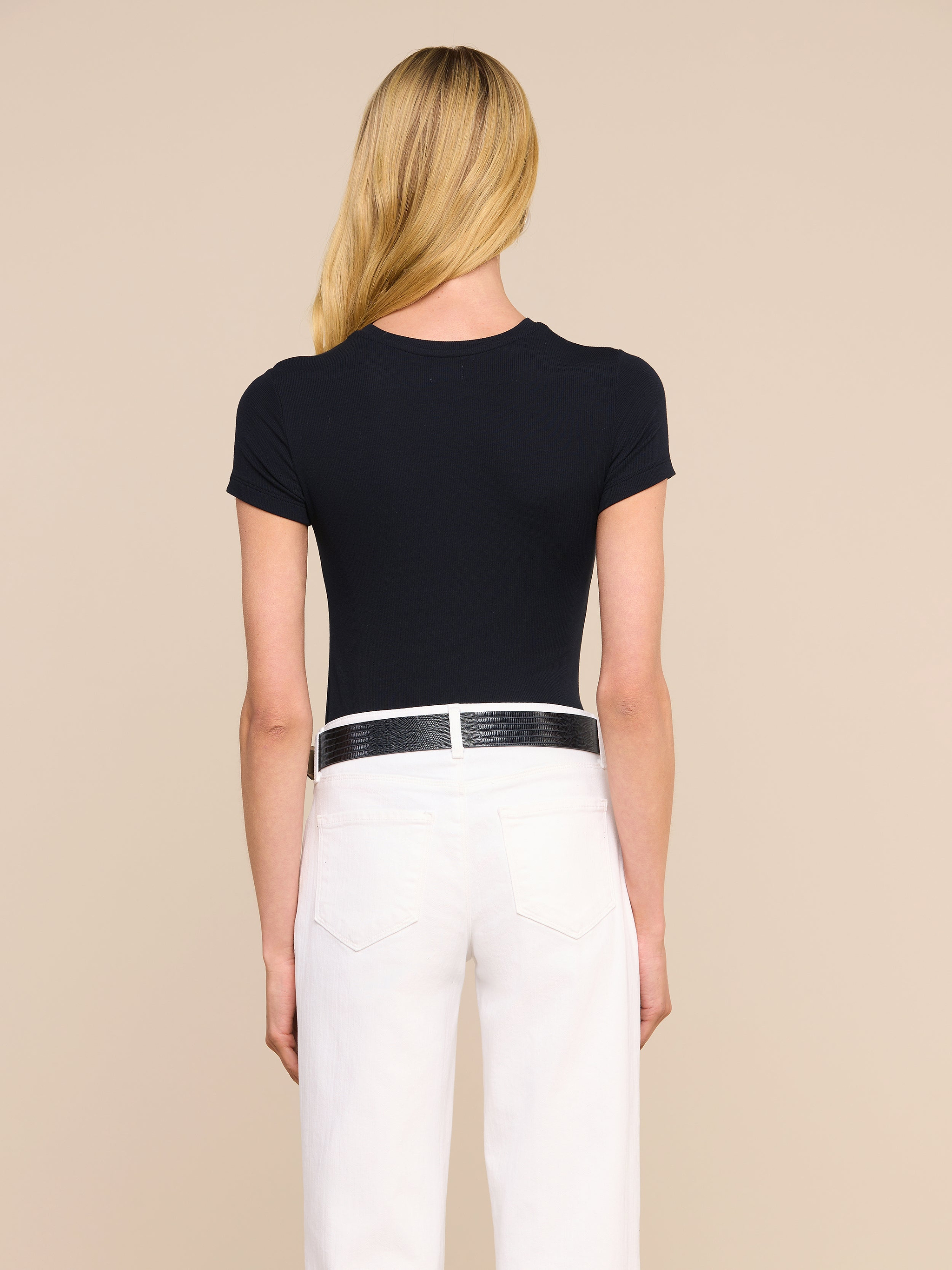 Una persona con lunghi capelli biondi è ritratta di spalle, con indosso la maglietta nera Ressi Micro-Rib, pantaloni bianchi e una cintura nera, su uno sfondo beige a tinta unita.
