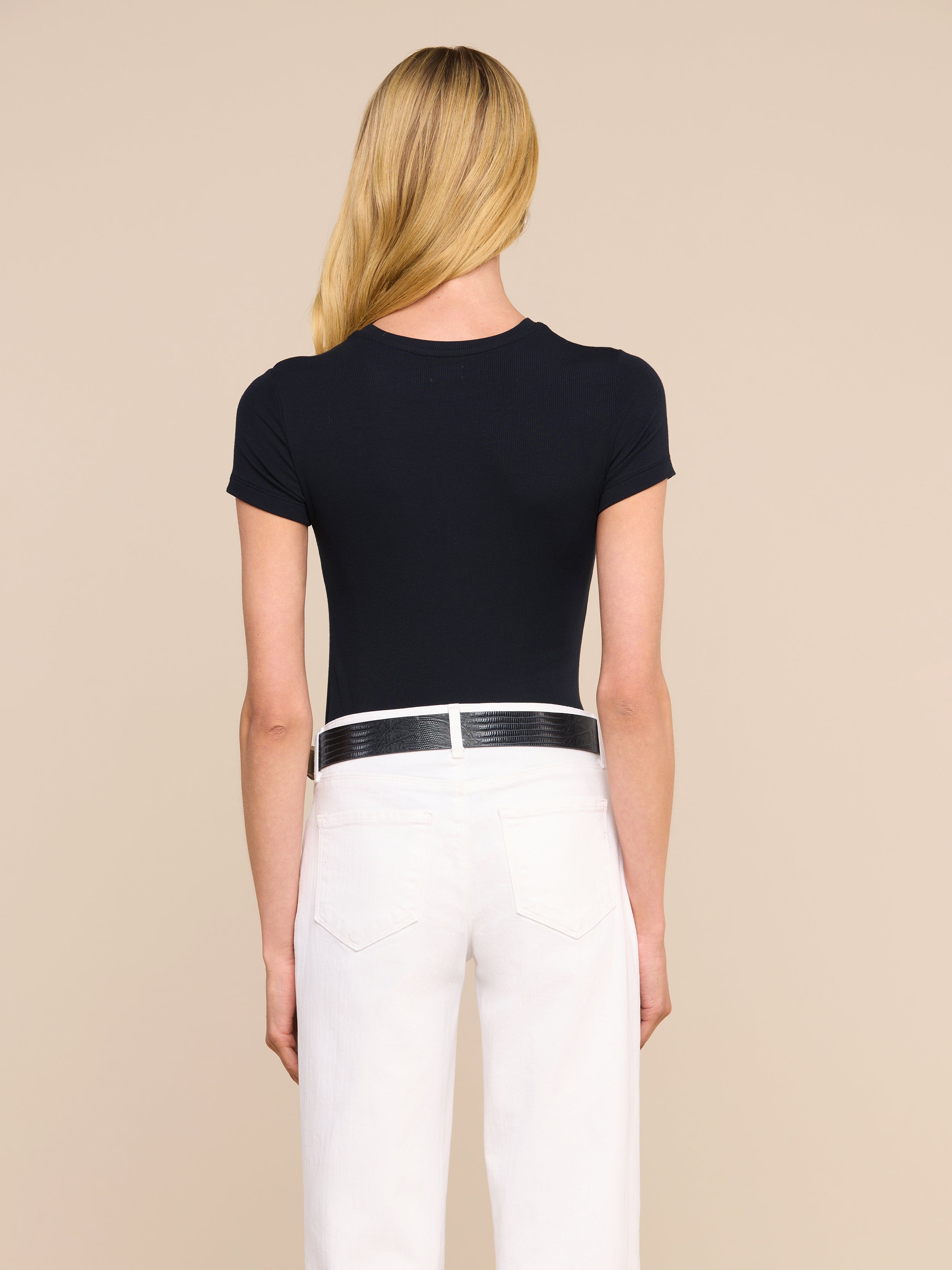 Una persona con lunghi capelli biondi è ritratta di spalle, con indosso la maglietta nera Ressi Micro-Rib, pantaloni bianchi e una cintura nera, su uno sfondo beige a tinta unita.