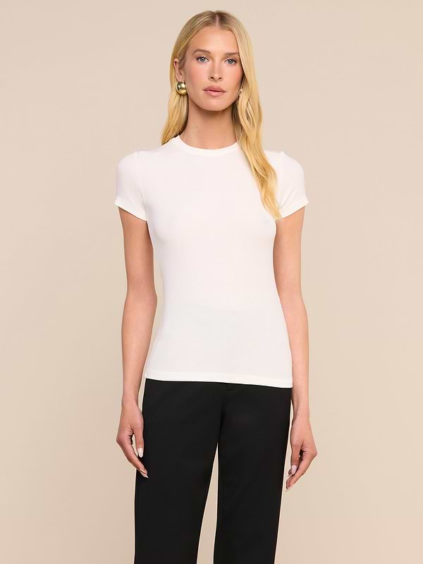 Une personne aux longs cheveux blonds, vêtue dun t-shirt Ressi Micro-Rib blanc et dun pantalon noir, se tient debout devant un fond beige uni, le regard tourné directement vers lappareil photo.