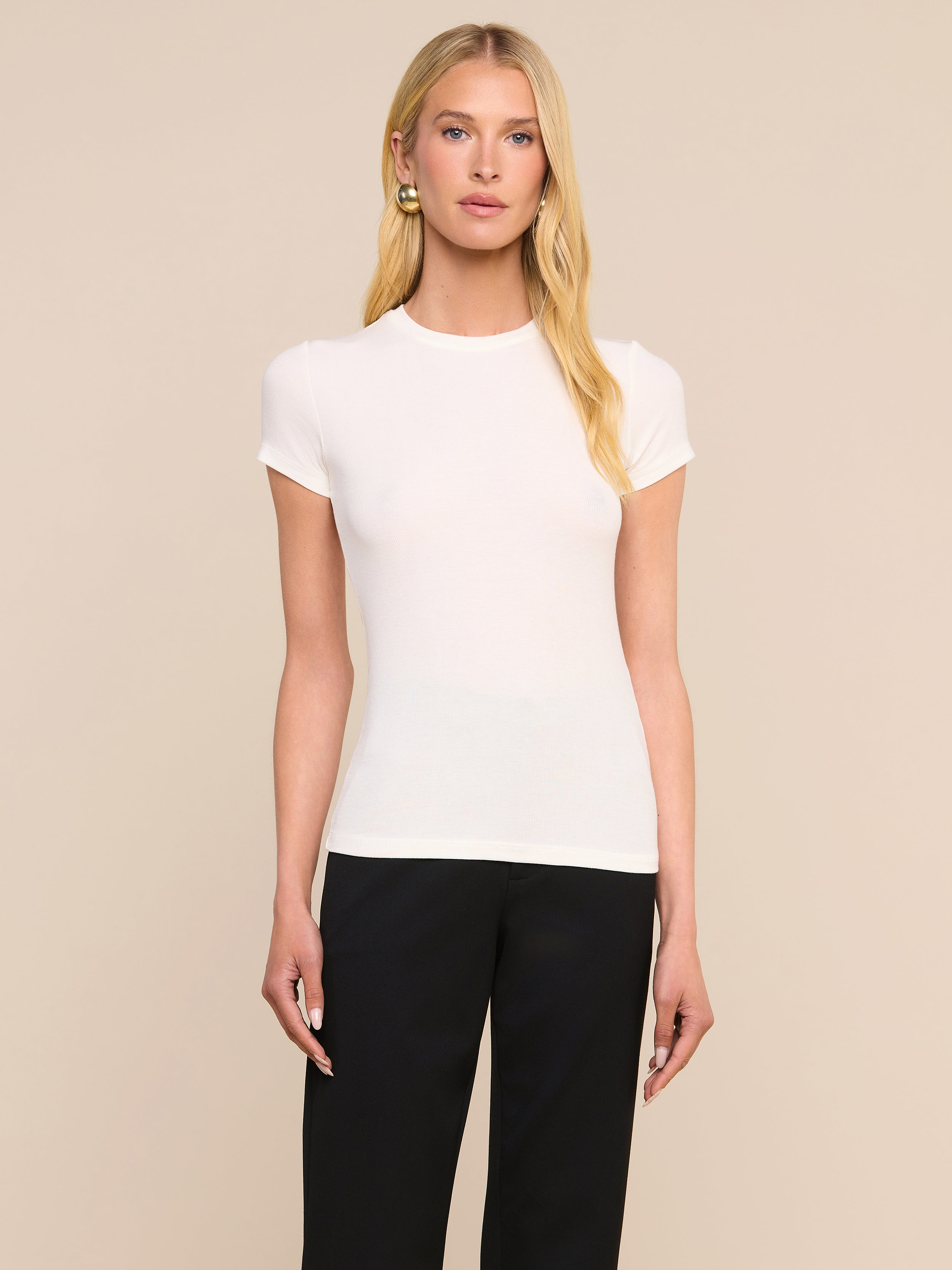 Une personne aux longs cheveux blonds, vêtue dun t-shirt Ressi Micro-Rib blanc et dun pantalon noir, se tient debout devant un fond beige uni, le regard tourné directement vers lappareil photo.