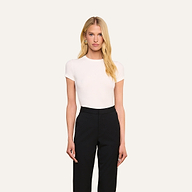 Une personne aux longs cheveux blonds se tient de face devant un fond beige uni, vêtue dun t-shirt blanc Ressi Micro-Rib et dun pantalon noir.