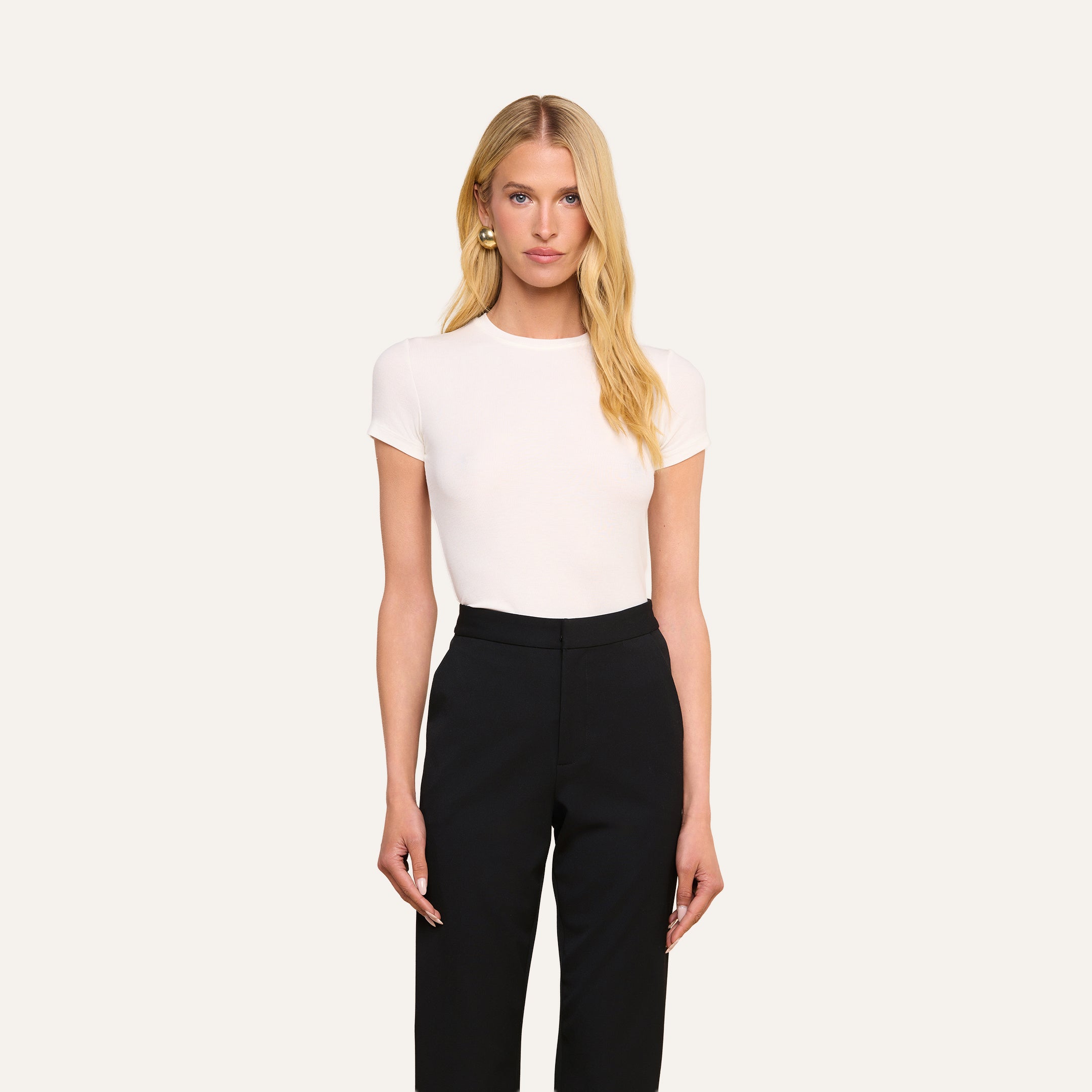 Une personne aux longs cheveux blonds se tient de face devant un fond beige uni, vêtue dun t-shirt blanc Ressi Micro-Rib et dun pantalon noir.