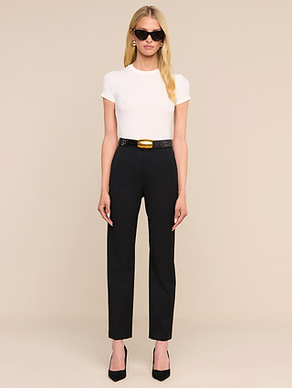 تقف امرأة ترتدي قميص «Ressi Micro-Rib Tee» باللون الأبيض، ونظارات شمسية سوداء، وسروالاً أسود مزوداً بحزام، وحذاءً بكعب عالٍ أسود، أمام خلفية بيج سادة.