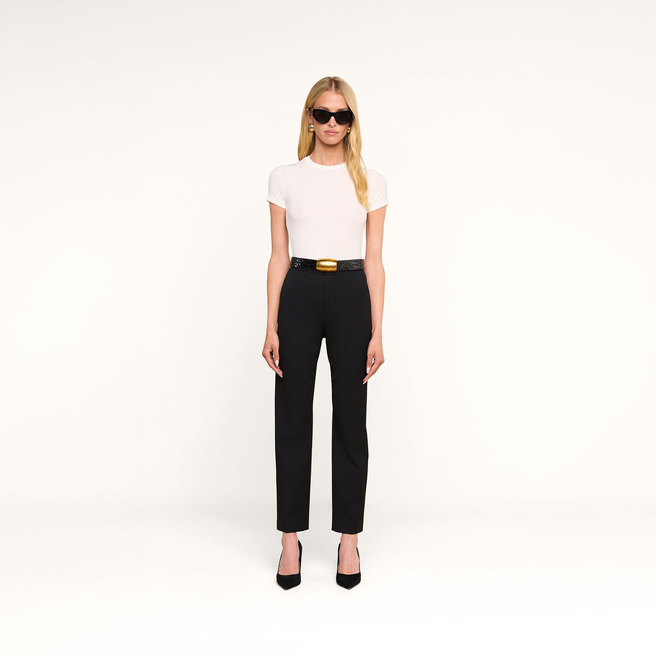 Une femme vêtue dun t-shirt « Ressi Micro-Rib » blanc, portant des lunettes de soleil noires, un pantalon noir à ceinture et des talons noirs, se tient debout devant un fond beige uni.