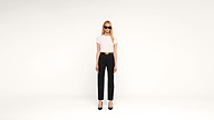 A woman in black sunglasses, a white top, black high-waisted pants, and black heels stands against a plain white background.