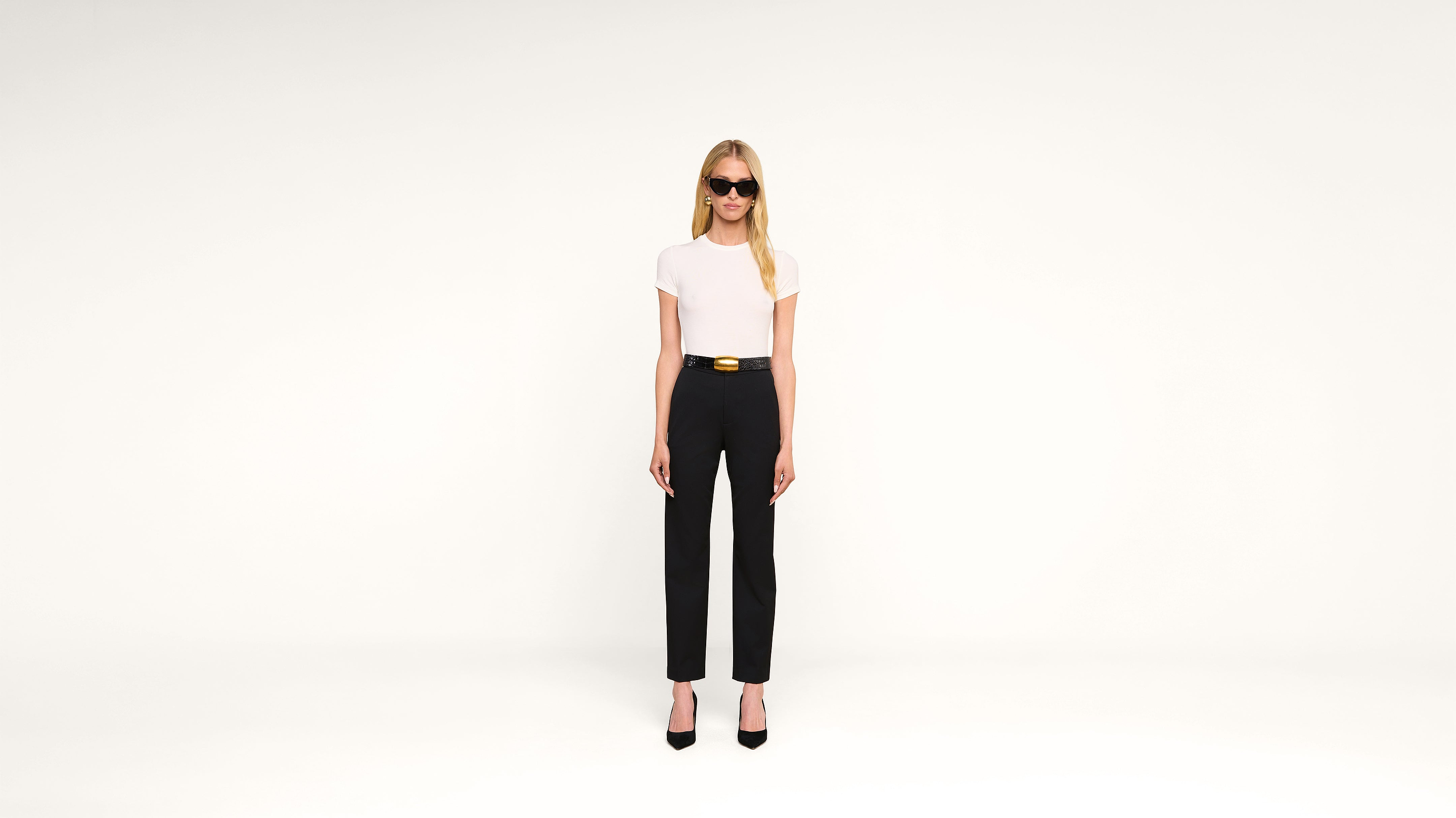 A woman in black sunglasses, a white top, black high-waisted pants, and black heels stands against a plain white background.