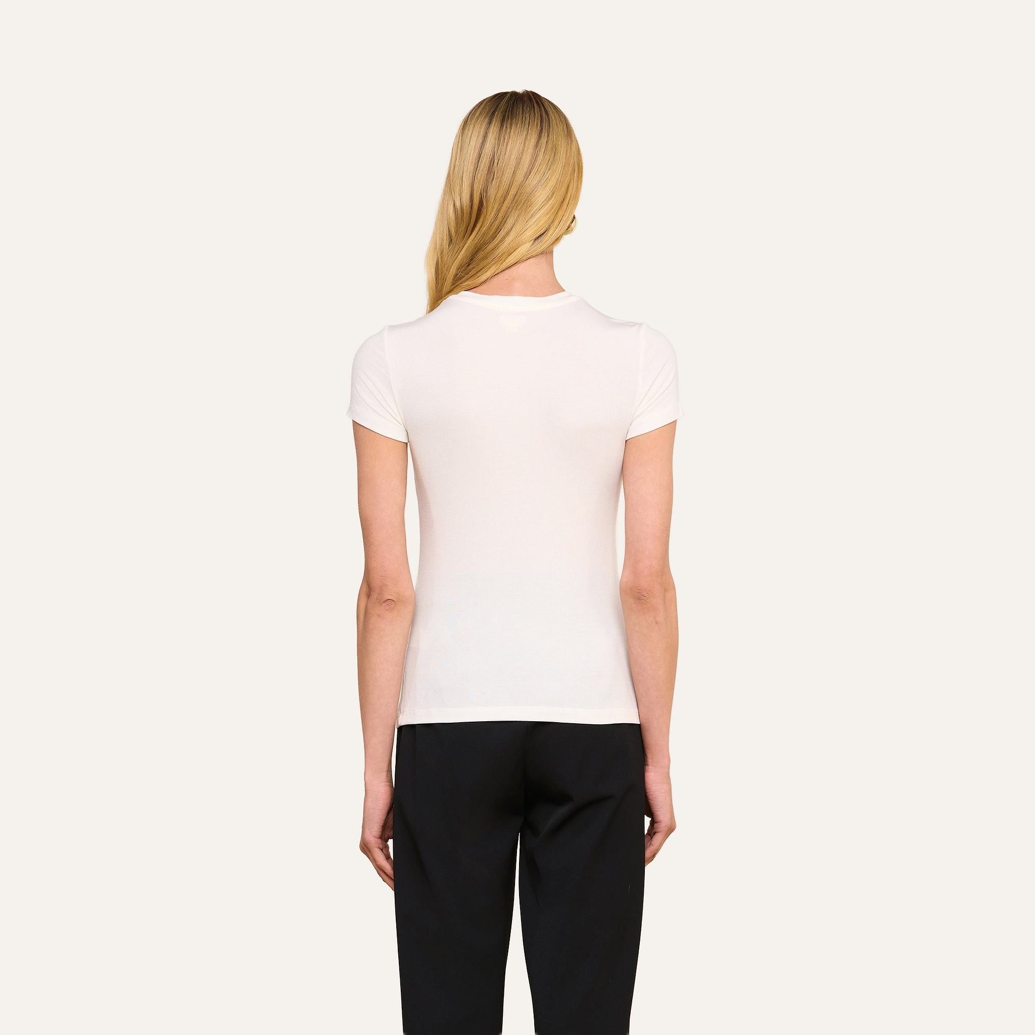 Une personne aux longs cheveux blonds est de dos, vêtue dun t-shirt blanc Ressi Micro-Rib ajusté à manches courtes et dun pantalon noir, sur un fond beige clair.