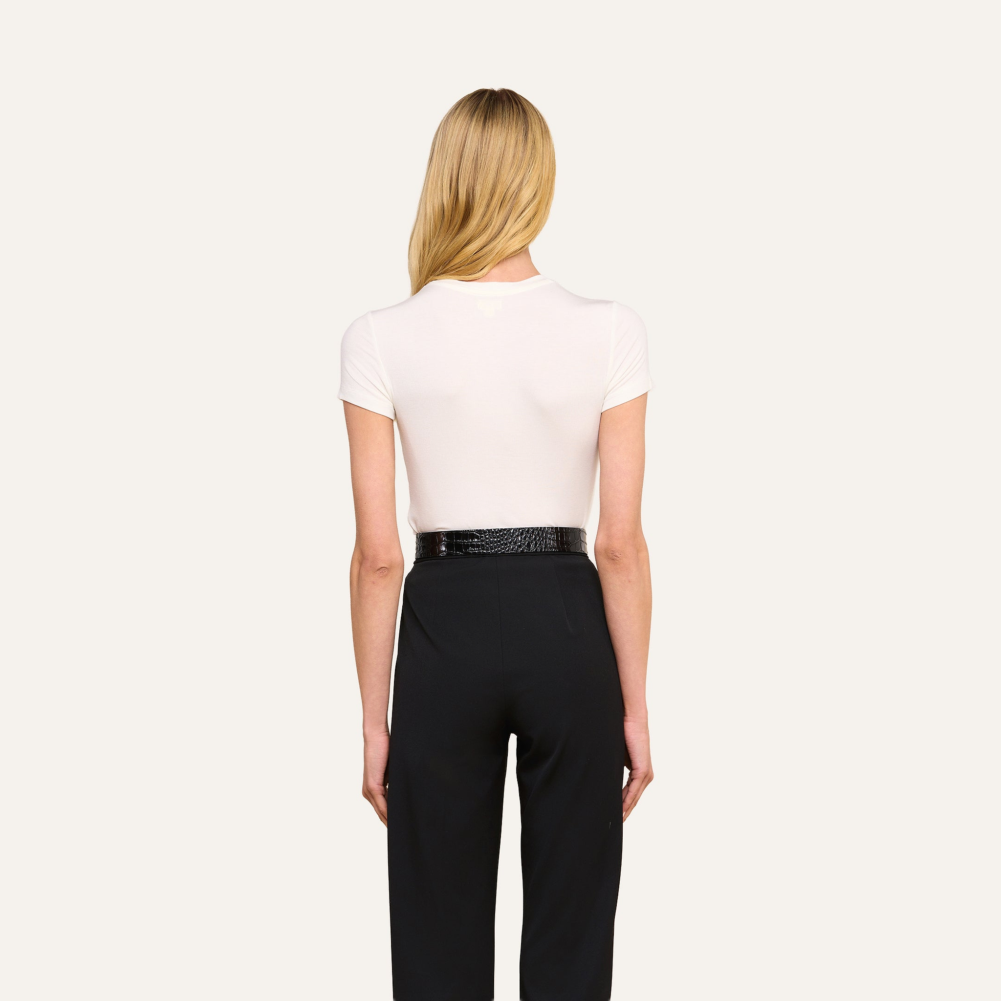 Une femme aux longs cheveux blonds se tient dos à la caméra. Elle porte le t-shirt « Ressi Micro-Rib » blanc, rentré dans un pantalon noir à taille haute, quelle porte avec une ceinture noire. Larrière-plan est dun beige uni.